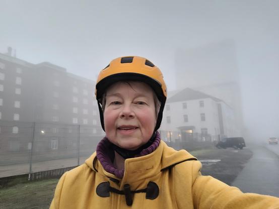 Eine ältere Frau im gelben Dufflecloat und mit einem dottergelben Fahrradhelm steht vor alten Gewerbe-Gebäuden, die im starken Nebel versinken.