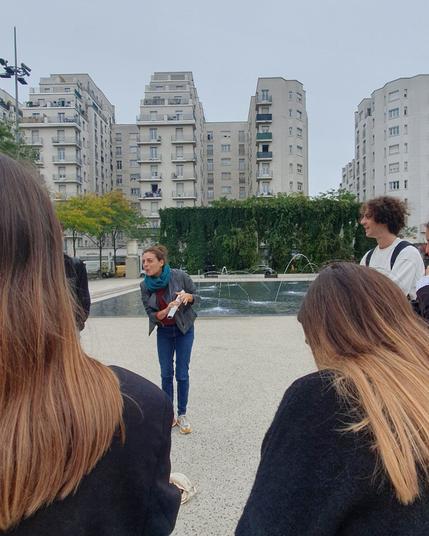 Photo de Chloé qui se penche en souriant dans une pose théâtrale, devant elle (au premier plan) des personnes lui font face. À l'arrière plan on voit un bassin avec des jets d'eau et de grands immeubles d'une dizaine d'étages avec des gradins au sommet.