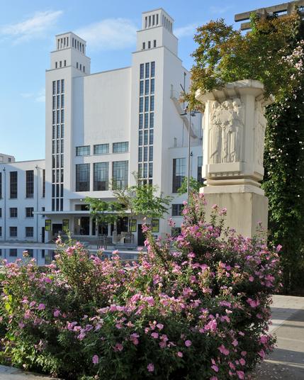 Photo du Théâtre (TNP) et sa façade art déco. Au premier plan il y a un buisson avec des fleurs roses en une statue art déco sur un pilier.