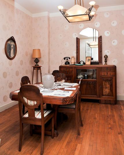 Photo d'un intérieur des années 30, c'est une salle à manger. Il y a une table et des chaises en bois et dans le fond de la pièce un buffet art déco avec un vieux téléphone posé dessus, quelques éléments décoratifs et un grand miroir. La tapisserie est d'époque avec de grosses fleurs beiges et le lustre art déco.