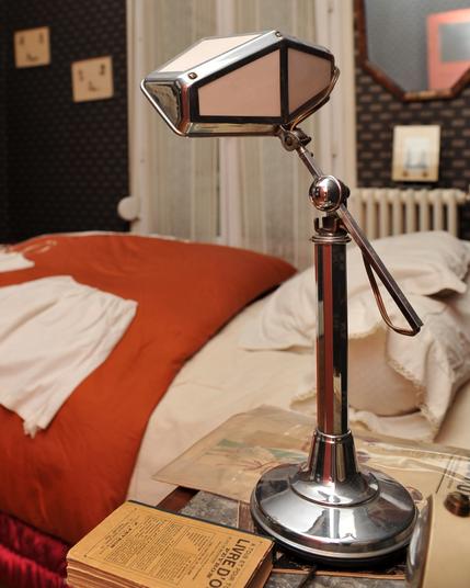 Au premier plan de la photo, une lampe des années 30 posée sur une table de chevet. On devine un livre et un journal assez anciens. Derrière, il y a un lit et dans le fond on devine le mur et sa tapisserie sombre, un miroir et d'autres éléments décoratifs.