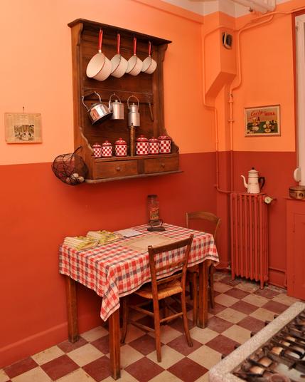Photo d'une cuisine des années 30 : une petite table recouverte d'une nappe à carreaux, un meuble en bois accroché au mur avec des pots anciens, des casseroles, et une vieille cafetières posées sur le radiateur.