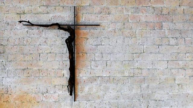 Vor einer Ziegelmauer befindet sich ein schwarzes dünnes Kreuz, dessen linke Seite aus Sicht des Betrachters länger ist als die rechte. Ein Teil eines dunklen Weinstocks ist an der unteren senkrechten und linken Seite des Kreuzes befestigt.