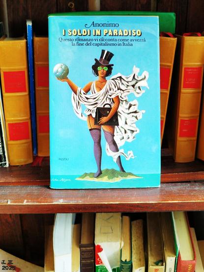 Blick in ein Buchtauschregal in Italien. Jemand hat ein Buch mit dem Titel "I soldi in paradiso" ("Geld im Paradies") in die Bildmitte gestellt. Dahinter eine Reihe von Bänden, die aussehen wie Teile einer Enzyklopädie. Im Regal darunter ein Sammelsurium von Buchoberteilen.