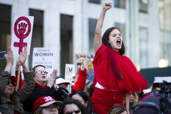 A vivacious lady with a clenched fist is shouting, behind her are a collection of protestors ones  of whom is is holding the traditional placard of Feminism