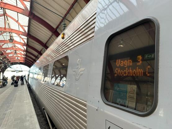 a view down a platform. A grey train is to the right. In the foreground, in a window, an amber LED destination board reads “Vagn 3 Stockholm C”