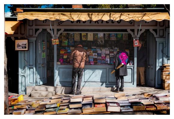 ### Motiv:
Das Bild zeigt einen kleinen Buchladen im Freien mit mehreren Regalen voller Bücher. Zwei Personen stehen vor dem Geschäft und betrachten die Auswahl, während eine dritte Person, die leicht versetzt im Hintergrund zu sehen ist, einen Blick auf die Bücher wirft. Auf dem Boden liegen zahlreiche Bücher in Reihen.

### Stimmung und Wirkung:
Die Atmosphäre vermittelt eine charmante, nostalgische Stimmung, die an entspannte Tage beim Stöbern in Büchern erinnert. Die warmen Lichtverhältnisse und die bunten Buchcover erzeugen einen einladenden und inspirierenden Eindruck, der zum Verweilen einlädt.

### Technische Details:
Das Bild zeigt kräftige Farben, insbesondere Blautöne des Ladens und lebendige Buchcover. Die Lichtverhältnisse sind hell, was die Details gut zur Geltung bringt. Die Komposition ist klar strukturiert, mit dem Buchladen im Mittelpunkt und den Menschen, die aktiv nach Büchern suchen, in einer harmonischen Anordnung.