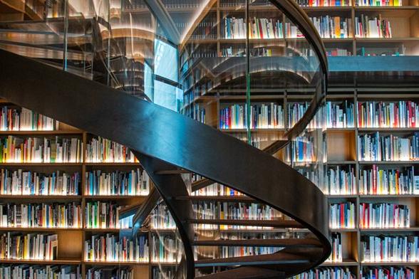 From bottom right to top left a spiral staircase with glass walls, a wide bronze edge at the bottom, bronze steps and a narrow edge at the top. Behind the staircase a bookcase full of books from top to bottom across the entire wall. The windows are reflected in the glass staircase.