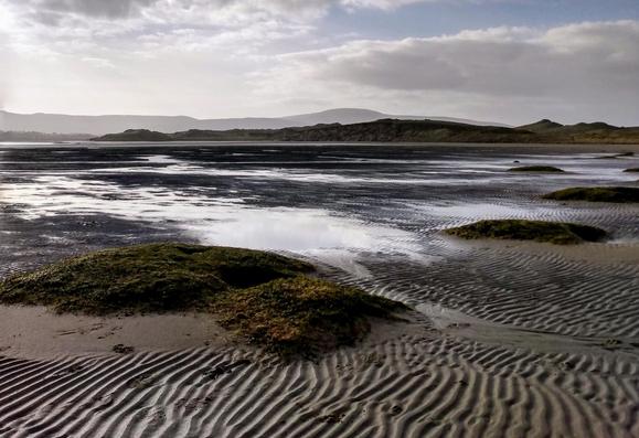 A bay at low tide. In front is the typical rippled, undulating sandy bottom, interspersed with puddles of seawater and small flat, overgrown sand hills. In the background, the opposite shore with dunes and the blurred Ox Mountains, set even further back, can be seen on the horizon. The sky is slightly cloudy.