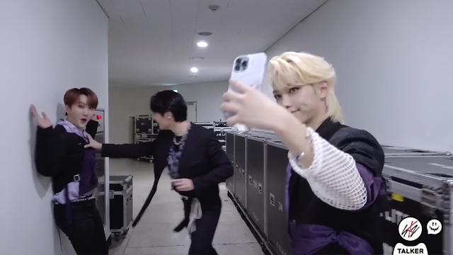 Felix taking a selfie. In the background, I.N pushes Changbin up against a wall with one arm. Changbin looks cutely at the camera and throws his arms up.