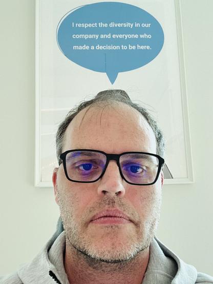 A man wearing glasses is staring at the camera. Above him is a speech bubble with the text: "I respect the diversity in our company and everyone who made a decision to be here." The background features a well-lit room with minimal decorations.