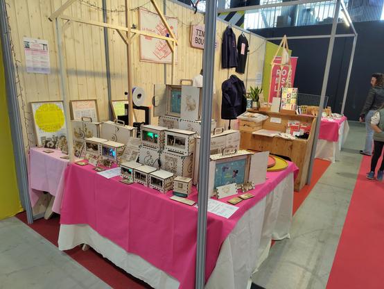 Stand du RoseLab à la foire de Toulouse 2025, centré sur la production de Denkodama : kamishibai et décorations lumineuses, tout en bois clair avec des gravures variées. Le tout est posé sur une nappe rose, une structure rappelle une forme de maison