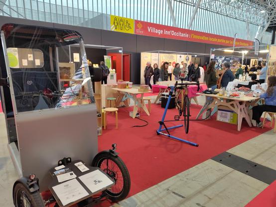 Le stand de démonstration du RoseLab à la foire de Toulouse 2025. Au premier plan à gauche, un véhicule intermédiaire (à mi chemin entre vélo et voiture) de maillon Mobility, puis un vélo à cadre en bois de chez Bois Roulant, un atelier coutureavrc trois machines à coudre, et au fond une maquette du RoseLab sur une table. En arrière plan, d'autres stands