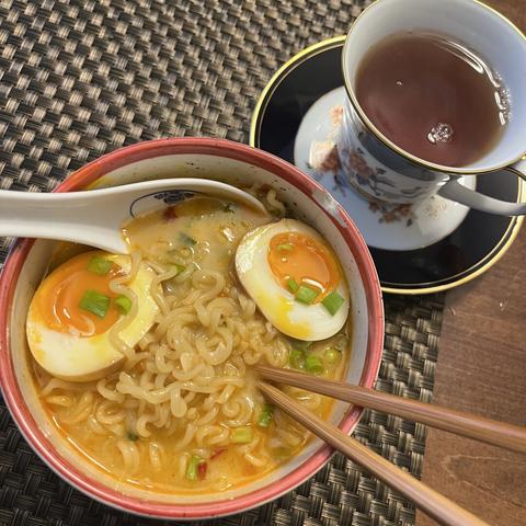 A bowl of ramen noodles with jammy soy sauce soaked eggs and green onions, accompanied by a cup of tea on a saucer.