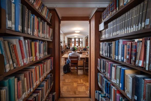 A library reading room is seen through a narrow passageway lined with books. Through the door are several tables with people seated at them.