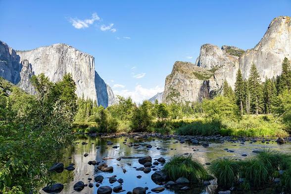 A scenic view of Yosemite National Park featuring the iconic granite cliffs of El Capitan, surrounded by lush trees, a reflective river, and a clear blue sky.