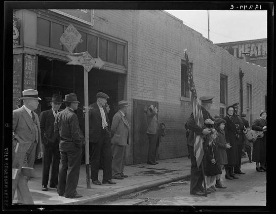 This black and white photograph captures a moment of community gathering on an urban street. A group of people, including men in suits and hats, women wearing coats, children with one holding onto the American flag pole, are lined up along the sidewalk next to a brick building labeled "Theatre." The crowd appears solemn or attentive as they face away from the camera towards what is not shown in the image.

There's a sign hanging on the side of the theater that reads "No Parking" and an arrow pointing upwards. Another street sign indicates it’s one-way with two streets intersecting at this point, but which direction each goes is unclear due to perspective. There are other signs for various businesses such as 'OKANES', 'WHISKEY BOURBON' indicating the presence of bars or liquor stores in close proximity.

The people's attire suggests a setting from several decades ago; men wear suits and hats typical of early 20th century fashion, while women don long coats indicative of colder weather. There’s also an individual with what appears to be musical instruments like violins held by the children which adds another layer of interest within this social scene.

The photograph is marked "3 HERSH" on one corner and "9-NOV21" at a top right, potentially indicating photographer or collection details along with date. The visual content portrays an everyday life scenario that could be reflective of cultural norms, societal interactions d [...]