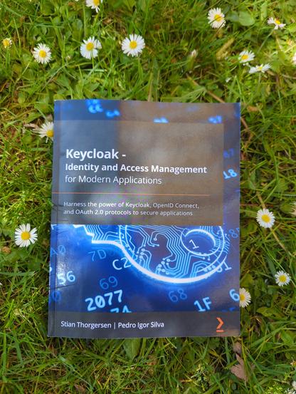A book lying in the grass with some dandelions. The book is mostly blue with some white numbers and graphics. The title, in a black box, is"Keycloak - Identity and Access management for modern applications" by Stian Thorgerson and Pedro Igor Silv"