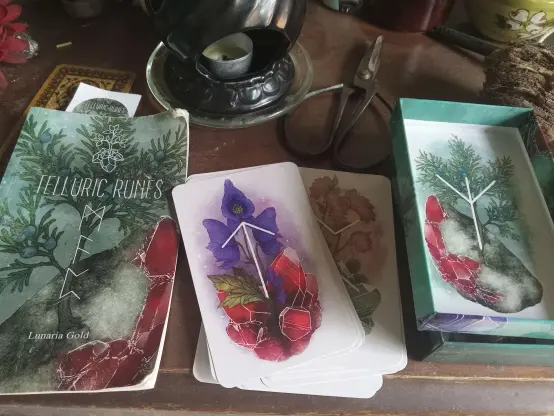 Flatlay photo of the telluric runes deck by lunaria gold. guidebook on the left, a pile of cards face up with tiwaz flanked by aconite plant and rubies in a luminous and vibrant art style in the middle. the open box with one card depicting algiz, flanked by juniper branches and tugtupite, on the right.