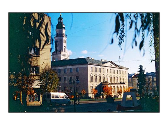 The image depicts a city square with a large, classical building in the center, featuring a symmetrical facade with multiple windows and a prominent clock tower with a green dome. The building is flanked by other structures, including a taller building to the left with a visible clock face. The square is surrounded by trees with autumn foliage, and a white van is parked on the street. The sky is clear and blue, suggesting a sunny day. The scene is framed by tree branches in the foreground, adding depth to the composition.