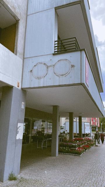 Hausecke mit Blumengeschäft, darüber der Schatten von etwas das früher einmal die brillenförmige Reklame Tafel für einen Optiker gewesen sein muss