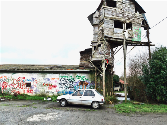 Photo d'extérieur d'un hangar tout tagué sur le bord duquel s'est bâtie une tour en bois très organique, sur des longs piliers
Devant, une vieille caisse blanche avec un chien dedans