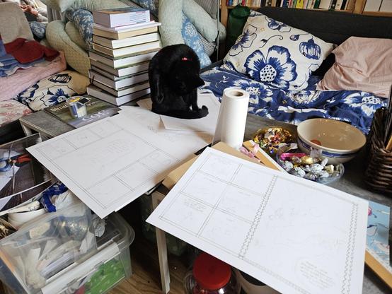 A chaos of unfinished comic pages, candy bowls, empty plates, trash, and plastic boxes with art supplies. Alea the Black Cat is taking a bath on top of one of my originals.