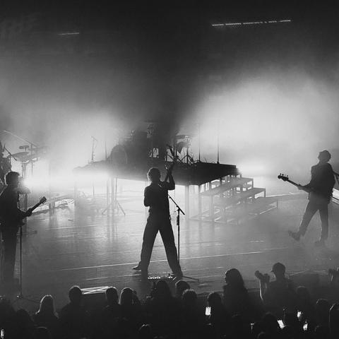This image shows a black and white scene of a band performing live on stage. The lighting creates a dramatic silhouette effect on the musicians. The audience is visible in the foreground, and there's stage equipment like microphones, guitars, and a drum set visible on stage.