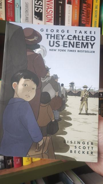 Cover to the graphic memoir They Called Us Enemy, showing a small boy in a line of adults heading into an internment camp, the child looking back nervously over his shoulder to the viewer