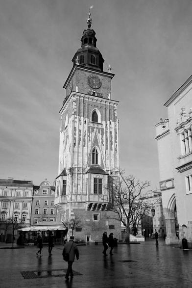 A black and white photograph shows the Town Hall Tower in Kraków, Poland, standing prominently in a paved square surrounded by other buildings and a few people walking. The sky is overcast.