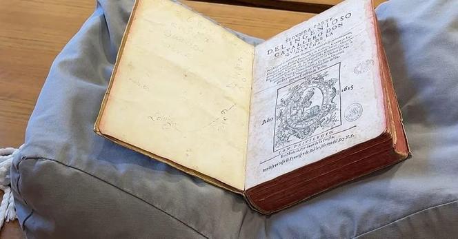 Ein offenes Vintage-Buch liegt auf einem grauen Kissen. Das Buch verfügt über kunstvolle Texte und Illustrationen auf seinen Seiten, mit einem Veröffentlichungsdatum von 1653 sichtbar. Das Cover zeigt den Verschleiß und zeigt sein Alter und seine Geschichte an.