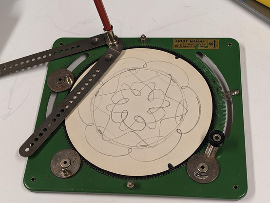 the hoot-nanny magic designer, a green metal device with a spinning platter in the center and several gears around it, with a pencil in a compass-like device for drawing on paper on the platter