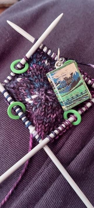 A small piece of knitting in a black, white, pink, and blue speckled yarn, on a set of 4 metal double pointed needles. Each needle has a small green hoop in the middle of the stitches and there is a tiny mock version of The Hobbit as a stitch marker that marks the start of a round.