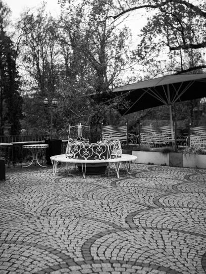 Black and white photo of a closed down outdoor cafe. Ornate wrought-iron furniture and large umbrellas are arranged on a cobblestone surface.  Central feature is a circular bench around a planter with an olive tree.