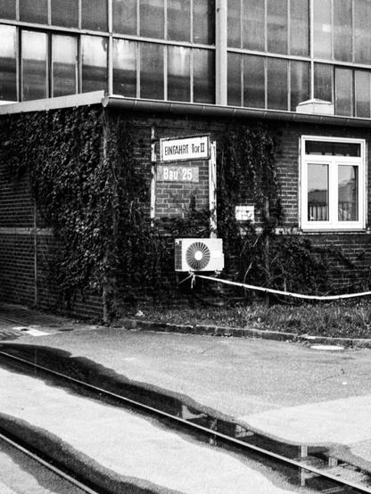 Black and white photo of an industrial building there is a brick annex in front of a steel and glass hall. Signs on the brick wall read "Bau 25" and "Einfahrt Tor II"