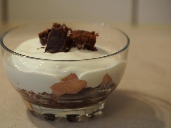 Das fertige Dessert in einem Glas: zu unterst eine Schicht Kuchenstücke, darüber das Rhabarberkompott, darauf der Joghurt und als Dekoration noch 2 oder 3 weitere Kuchenstückchen.