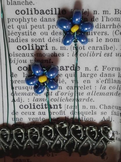 Forget-me-nots made with tiny glass rocailles on green wire in front of an old dictionnary page. The glass beads for a flower: 5 blue lustered ones and one small yellow one in the middle.