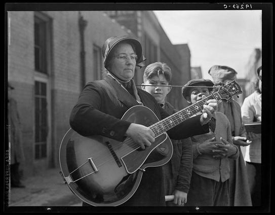 The black and white photograph depicts a group of people standing on what appears to be an urban street. In the foreground, there is a woman wearing glasses, a dark coat with buttons, and a hat adorned with a decorative bow or ribbon in her hair. She holds a guitar firmly with both hands as she looks directly at the camera, suggesting that she may have been playing music.

Behind her stands another individual who seems to be singing into an unseen microphone, possibly contributing vocals for a street performance. This person wears a coat and hat similar to the woman's attire but is not holding any musical instrument in their visible hand.

In addition to these two figures, there are several other people present. One young boy appears in the background on the right side of the image; he looks towards the camera with an expression that could be interpreted as focused or contemplative. Another person can also be seen standing slightly behind and to the left of him.

The environment around them suggests a busy street setting, possibly during a community event or gathering where people come together for entertainment purposes. The buildings in the background have windows, and there is no clear indication of any specific location beyond urban surroundings. This image evokes an atmosphere reminiscent of early 20th-century America, as suggested by its black-and-white nature and street performances which were popular during that  [...]