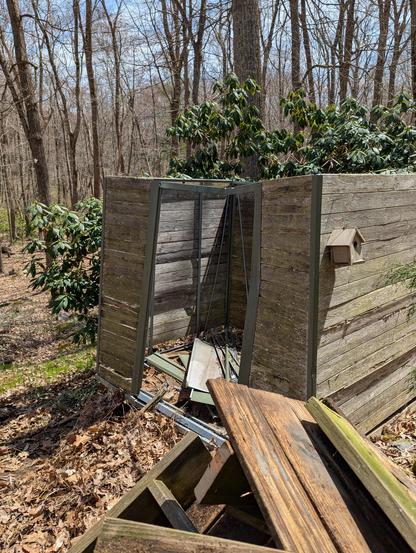 A partially built small building that was never completed. It is next to be dismantled.  Behind it is a large green rhododendron. There's a small birdhouse on one side of the structure. In front is a pile of deck pieces.