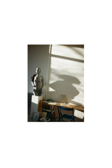 A photo, taken with an analog camera, of an interior scene of an art studio. On the left is the sculpture of a woman, which casts a shadow above the artist's desk.