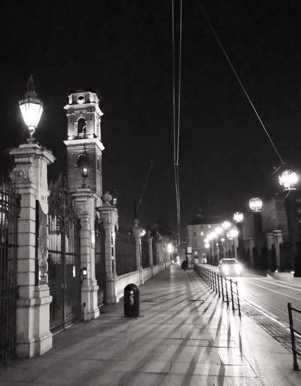 The photo shows a black and white nighttime street scene. A pathway with ornate lampposts and a tall clock tower to the left. There are streetlights casting long shadows, and a road with a moving car on the right.