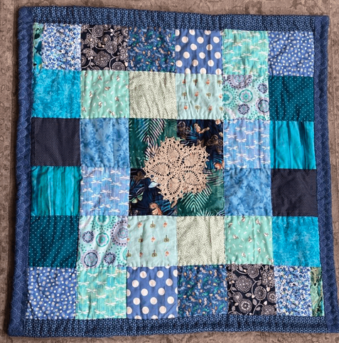 A simple quilt made from 36 squares arranged in a 6x6 square; the squares are all in blue and green and turquoise prints; there is a blue printed border; there is a five-pointed flower-shaped scrap of antique lace appliqued in the center, over darker squares.