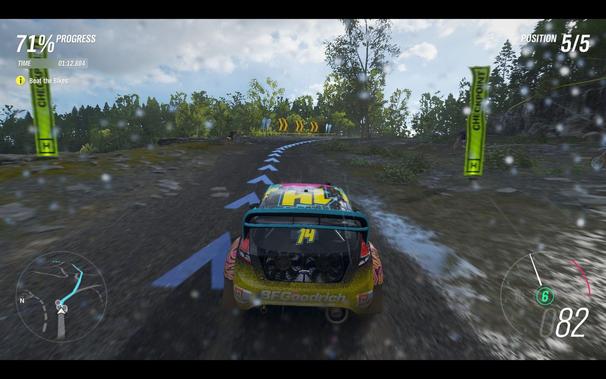 A dynamic racing scene set in a rain-soaked forest clearing. The players car, which is yellow and black, with the number 82 on it, is leading a race. Other cars are visible behind the players, indicating competition. The game interface shows a blue arrow pointing to the right, suggesting a path forward or objective.