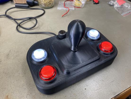 Fully assembled 3d printed joystick on my workbench. It has four fire buttons (2 white and 2 red) and a potentiometer to adjust the autofire speed. It’s made out of black plastic.