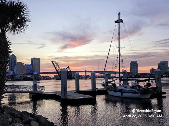Early morning well before sunrise, overlooking a vast river and the downtown skyline with bridges. In the foreground a sailboat is tied up at a small marina. The city skyline and bridges are illuminated by pastel colors rising from the horizon in advance of sunrise.