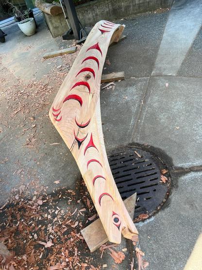 High-angle, medium shot of a light-colored, sculpted wooden fish placed diagonally across a gray concrete surface. The fish is carved with stylized red crescent shapes along its top edge, some filled with black. It rests on wood scraps amid wood shavings, with a dark, circular storm drain and cover positioned on the right side of the frame. The composition emphasizes texture contrast between the wood grain. The wood carvings contrast against the concrete. The sculpture is partially in shadow with good lighting on the overall form.