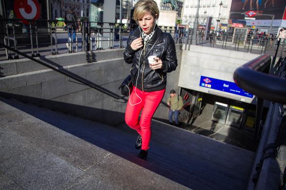 A candid street photograph taken in Madrid, Spain, showing a woman with short blonde hair wearing a black leather jacket and bright red trousers, walking up the steps from the Sol metro station while holding a coffee cup and listening to earphones; the blue and white "Sol" metro sign is visible in the background along with another person ascending the stairs.