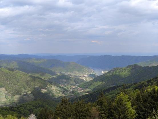 Blick ins write weite tiefe Tal hinab. Auf beiden Seiten sind Berge mit Wald zu sehen. Unten mehrere Ortschaften und am Ende des Tals ein Fluss. Himmel ist bewölkt.