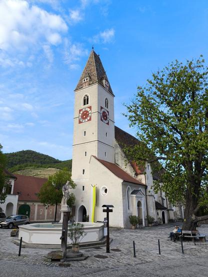 Ein Kirchturm in der Nahaufnahme. Davor am Kirchplatz ein Springbrunnen und daneben ein großer Baum mit Bank rundherum, auf der zwei Personen sitzen. Blauer Himmel.