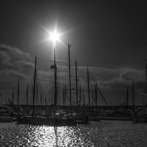 Man sie die Silhouetten von Segelschiffen am Anleger. Oberhalb vom größen Mast ist ein Sonnenstern zu sehen. Das Foto ist in Schwarzweiß.